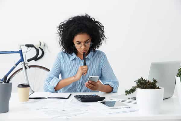 Frustrated young female entrepreneur in formal shirt and eyewear having a problem while working on financial report in office, reading information on mobile phone screen with perplexed expression